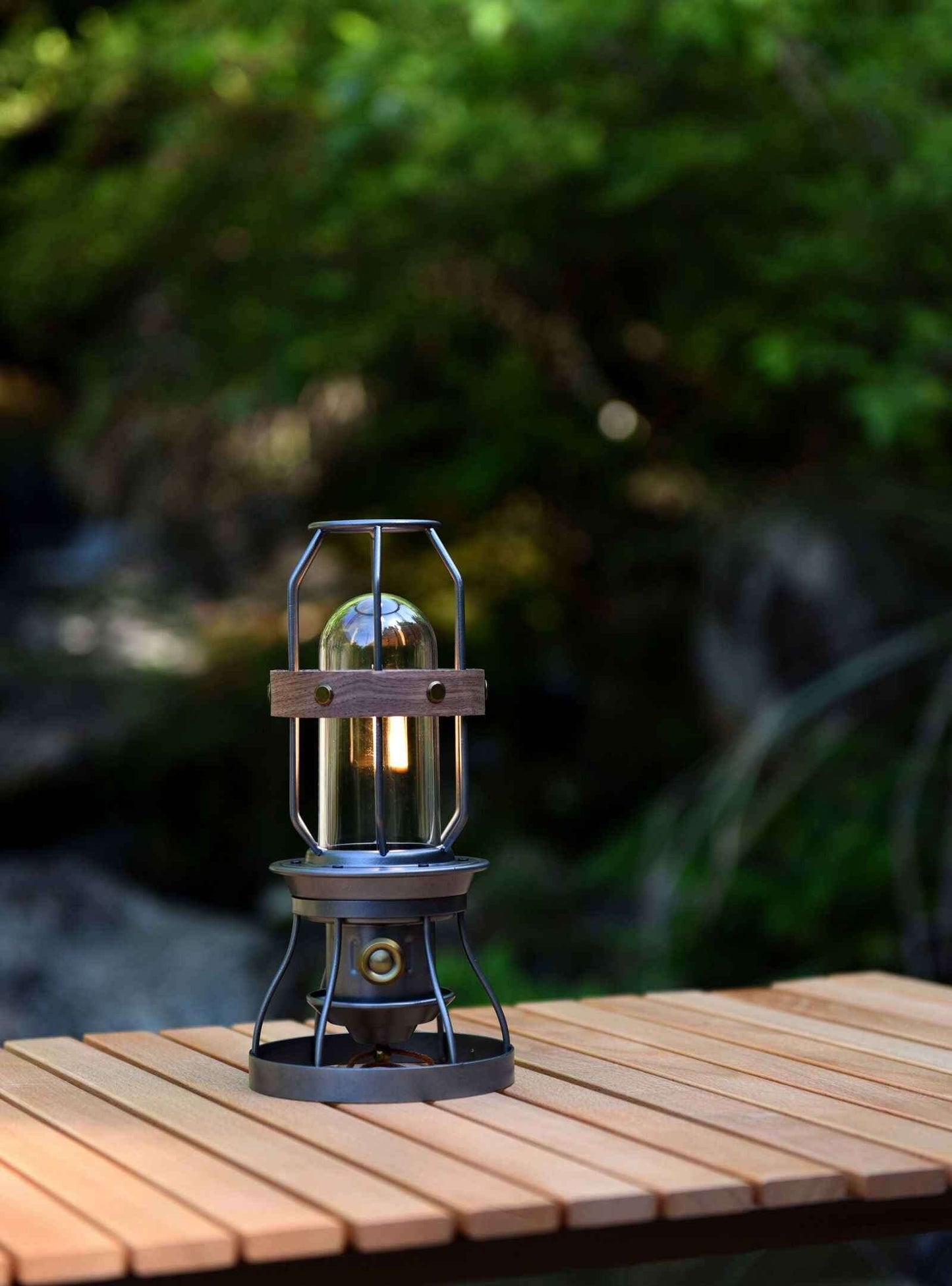 Vintage grey camping lantern on a wooden table outdoors surrounded by greenery