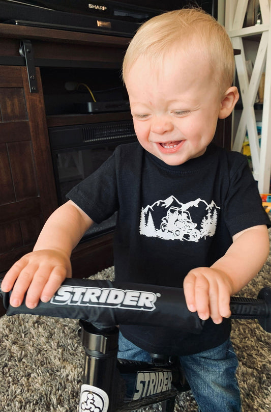 Toddler wearing a Dirt kids t-shirt Canada while happily riding a Strider bike indoors