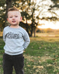 Toddler wearing a gray Happy Camper t-shirt outdoors in a grassy field at sunset